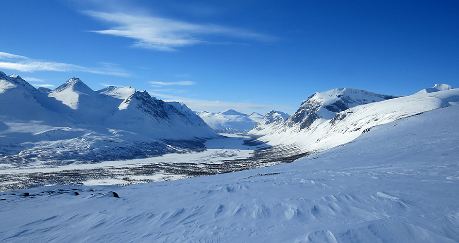 Vy över Rapadalen. De snöklädda fjällen tornar upp sig mot den blåa himlen runt dalen.