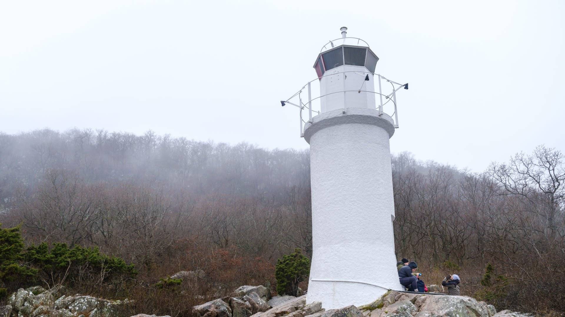 En liten vit fyr står på klippstranden vid bergets utsida. Några besökare sitter och fikar vid ingången. Stenshuvuds nationalpark. 