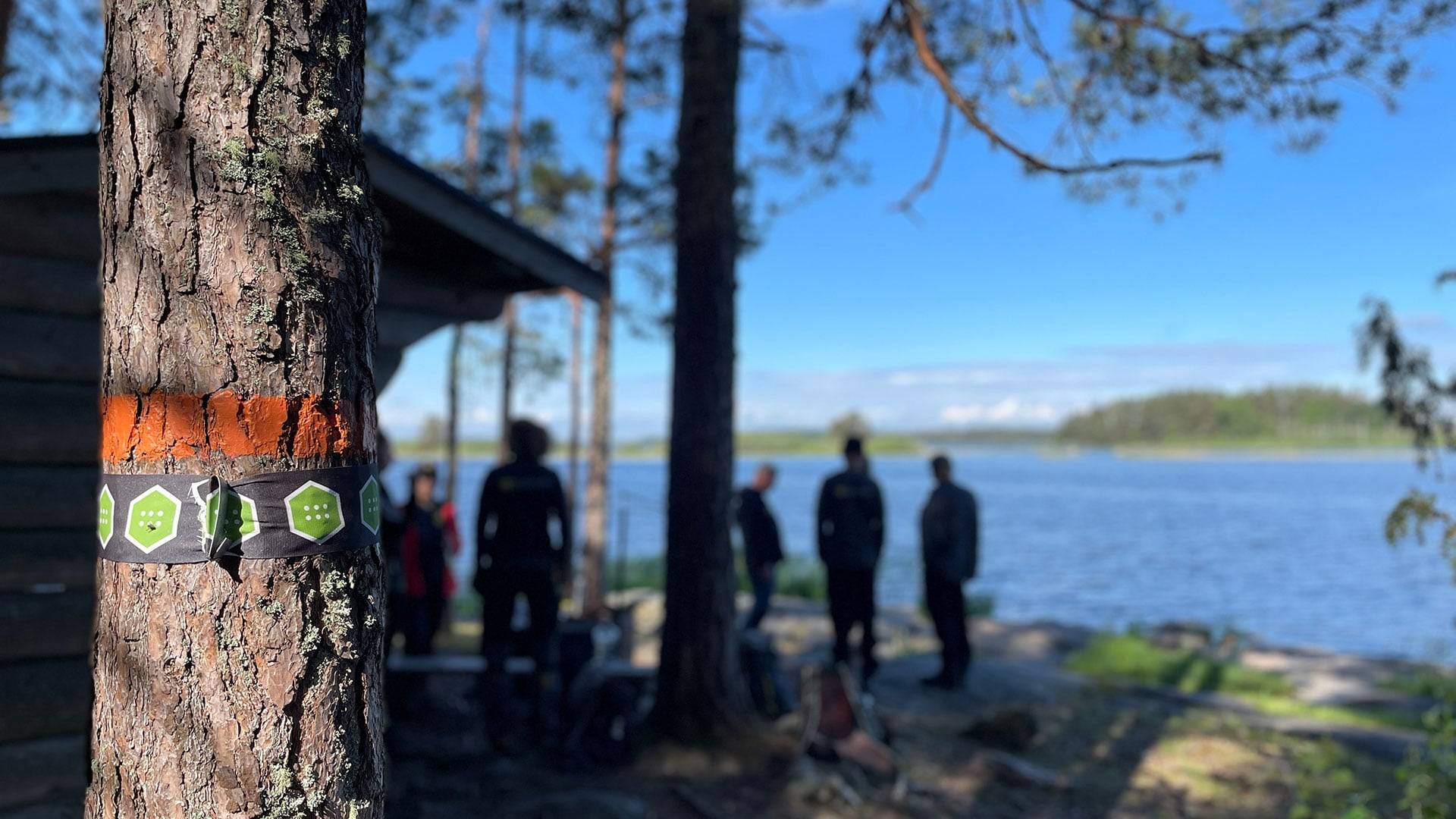 Markerad vandringsled, Skekarsboslingan i Färnebofjärdens nationalpark, på träd vid sjöstrand, med personer och byggnad i bakgrunden.