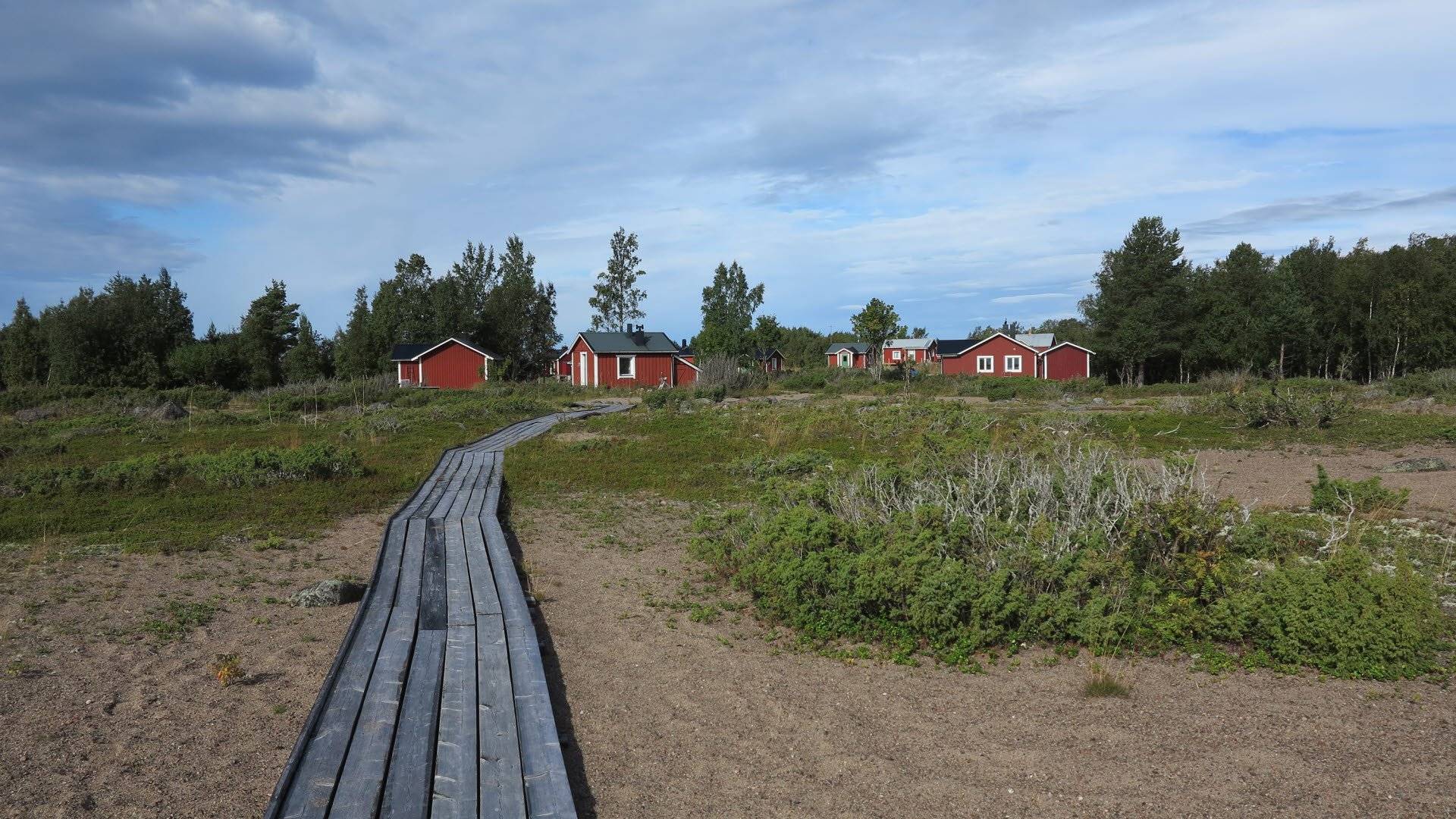 Träspång som leder fram till flera röda stugor, omgivna av träd och grönska. Haparanda skärgårds nationalpark, Norrbotten län, Sverige.