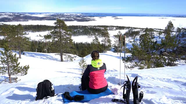 Foto av en person som sitter på ett liggunderlag i snön på en utsiktshöjd. Bredvid står en ryggsäck och några snöskor.