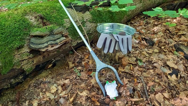 Skräpplockare, handskar och metallhink på marken i lövskog.