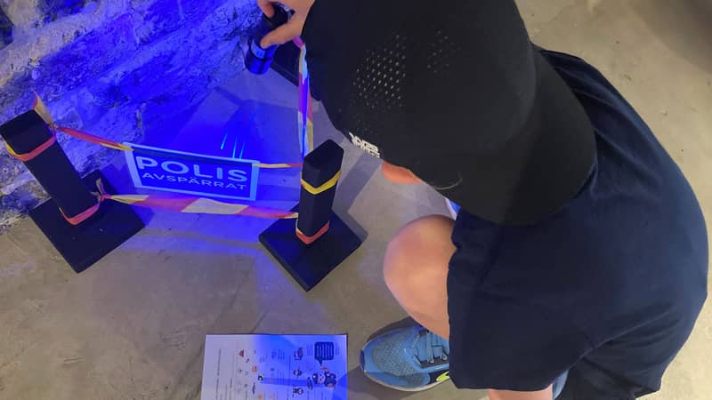 Länsstyrelsen Gävleborg Ett barn sitter på huk och lyser med UV-lampa innanför en låtsas-polisavspärrning på naturum Färnebofjärden.