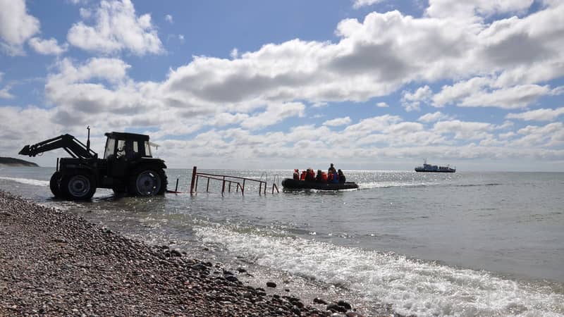 Magnus Lepschi Personer läktras mellan strand och fartyg i gummibåt, med traktor vid strandkanten.