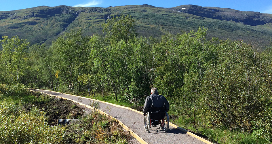 Man in a wheelchair on the trail with green mountains in the background.
