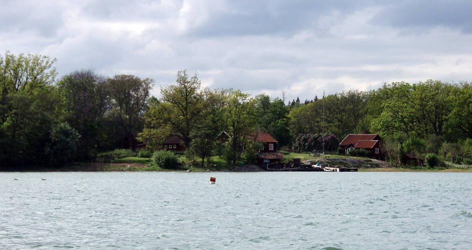 View from the water. Several crofts, a jetty and trees can be seen on land.