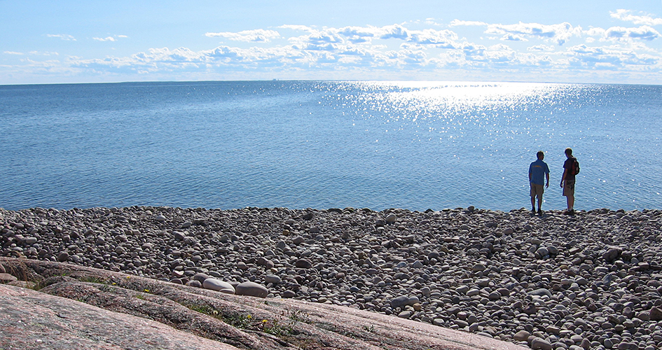 Strand av rundslipade stenar och glittrande hav. På stranden står två personer.