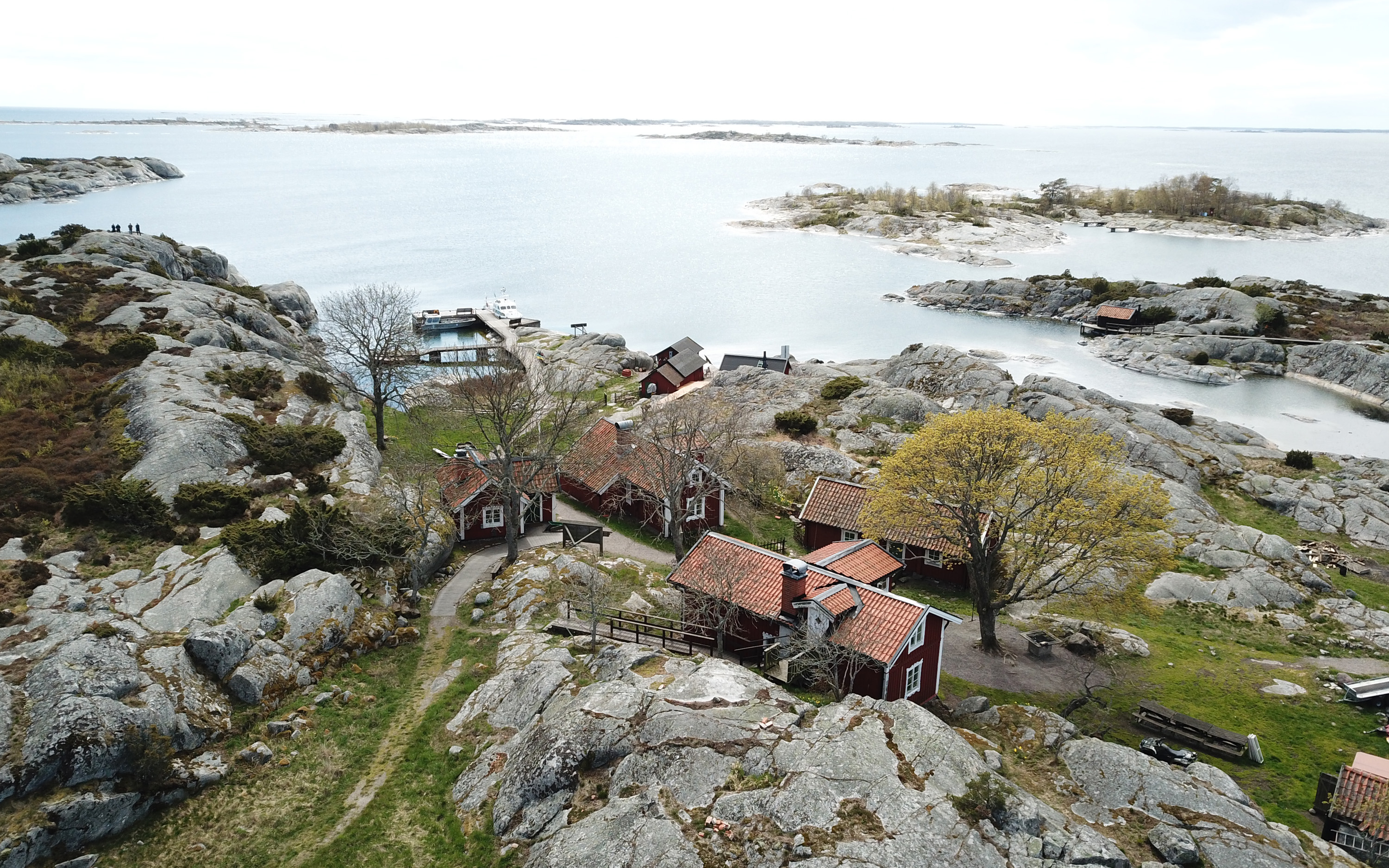 Flera hus bland klippor och gräs. I bakgrunden syns en havsvik. Bullerö, Nämdöskärgården.