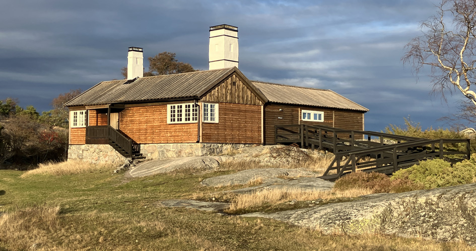 Stuga omgiven av gräs och klippor. En ramp går upp till stugan.