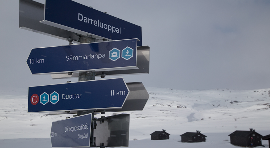 Hänvisningsskyltar i Padjelanta Badjelannda nationalpark med snötäckta stugor och grå himmel bakom.