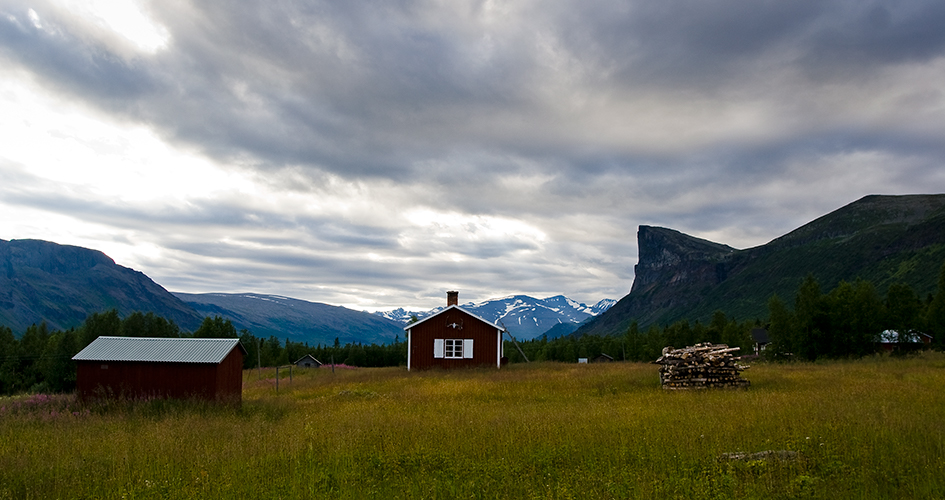 Två röda turiststugor i Aktse. I förgrunden en grän äng, i bakgrunden fjäll mot grå och molnig himmel.