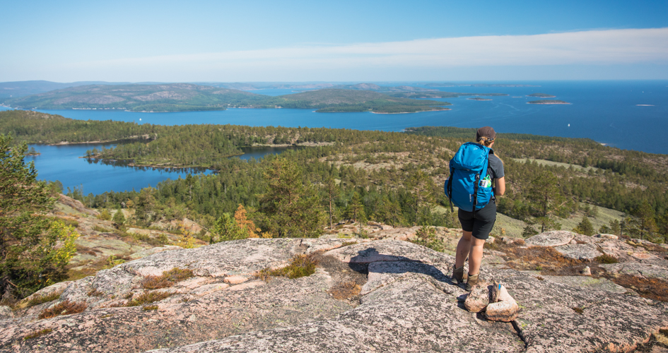 En tjej med stor ryggsäck som står på en klippa framför en utsikt ut över skog, tjärnar och havet.