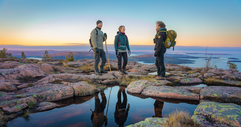 Three people standing and talking on a cliff with the ocean and a coloful sunset in the background.