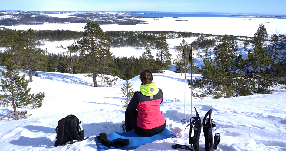 A person som sitter framför en utsikt ner över skog och isklätt hav. Det är vinter och mycket snö. Hon har med sig en ryggsäck och snöskor.