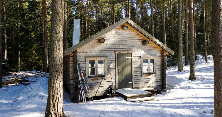 Timrad stuga omgiven av höga tallar och snö. Det står skidor och stavar utanför dörren.