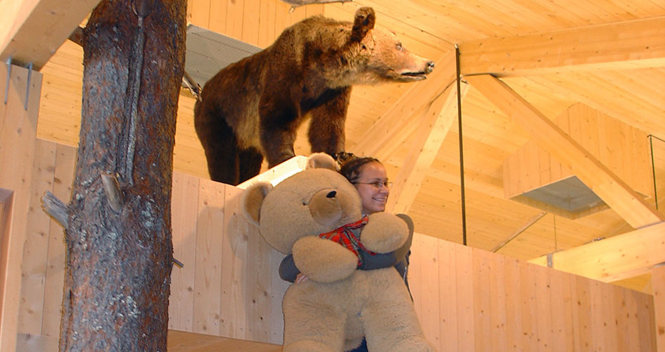 En kvinna kramar en stor teddybjörn inne i naturumhuset. Ovanför henne ses en uppstoppad brunbjörn.