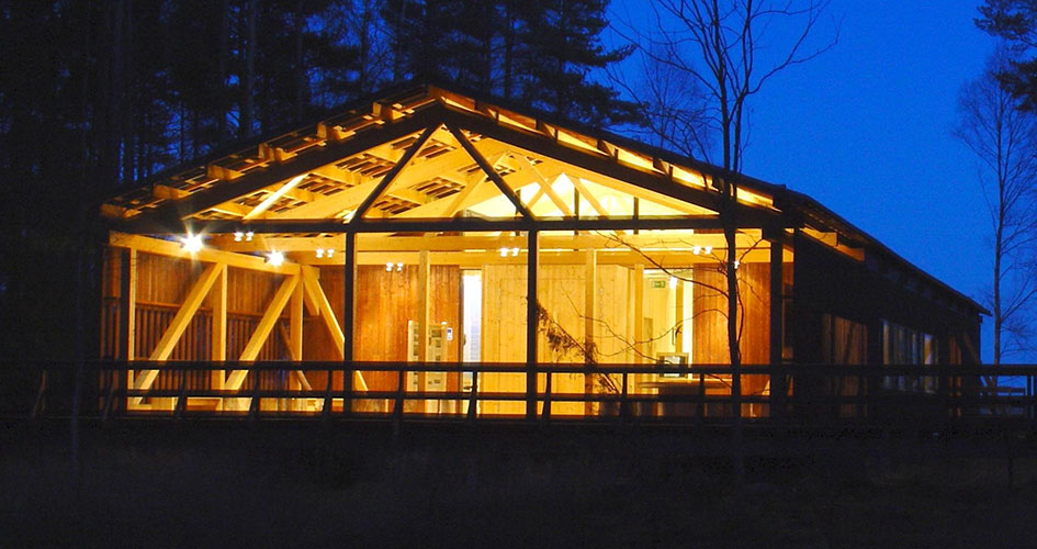 Naturum nattetid. En träbyggnad med siluett av en skog runtom. Ljusen lyser i orange från huset.