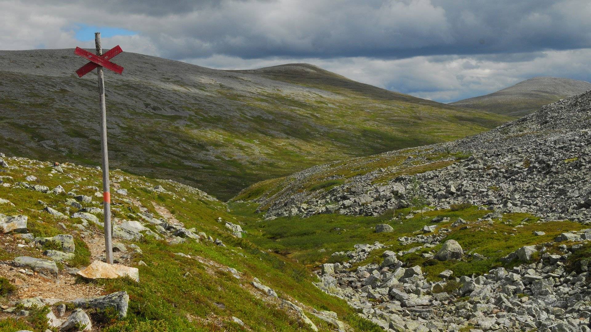 Fjällandskap med stenig mark och en röd ledkryssmarkering i förgrunden, gröna sluttningar och grå fjälltoppar under molnig himmel i Sonfjällets nationalpark.