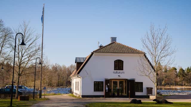 Andreas Lindahl / Naturvårdsverket Byggnaden som rymmer naturum ligger vid en grusad gångväg omgiven av träd och öppet landskap under en klar blå himmel.