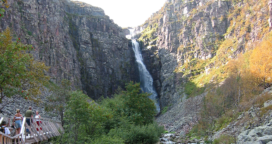 Ett forsande vattenfall, till vänster går människor på en gångbro i trä.