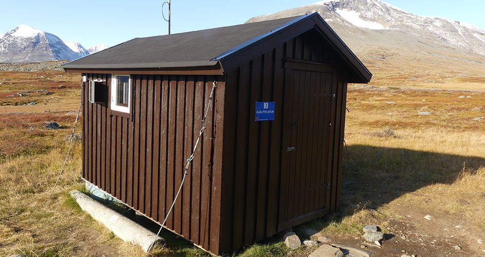 Liten brun stuga med snökladda fjall mot blå himmel i bakgrunden.