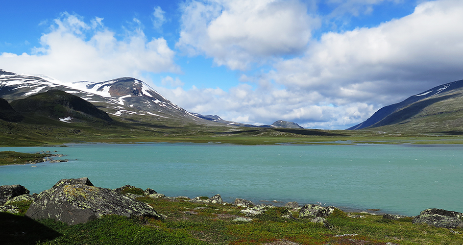 Vy över Guhkesvágge dalgång. En blågrön sjö ligger framför stora fjäll i bakgrunden. Sarek Nationalpark 