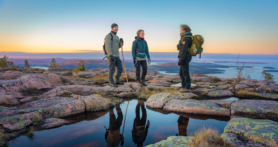 Tre personer som står och pratar på några klippor. Bakom dem syns havet och en vacker solnedgång.