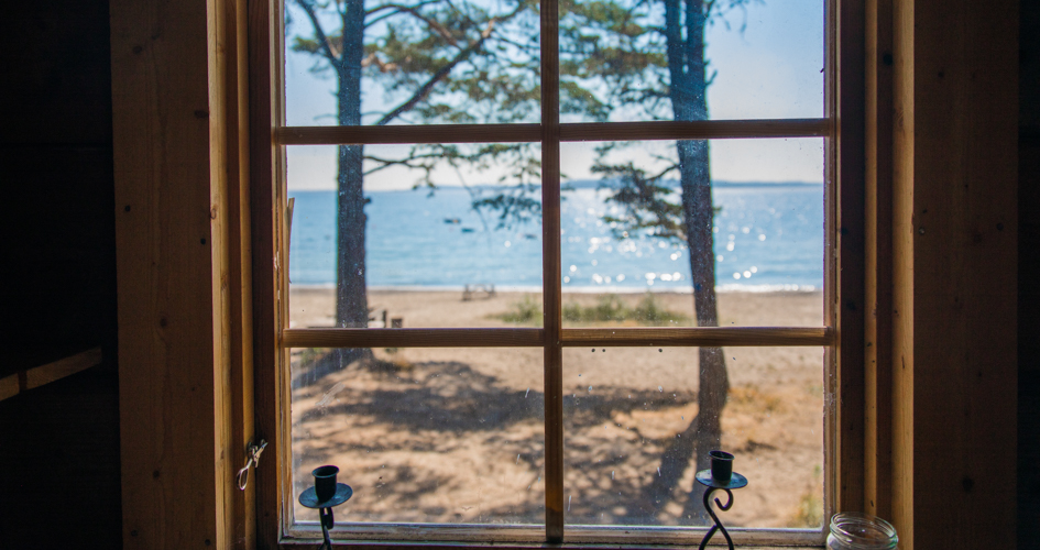 Utsikt genom ett fönster ut mot en sandstrand och havet.