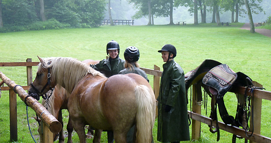 Två hästar och tre tjejer med ridhjälm och regnkläder står vid hästuppställningsplatsen Skaralid. Bakom inhägnaden ligger och grön gräsmatta som omsluts av skog.