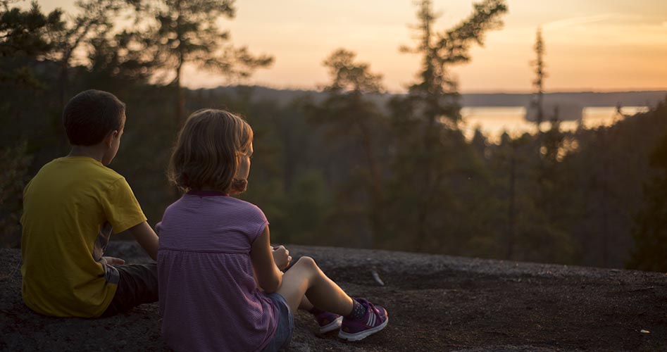 Två barn sitter på ett berg och blickar ut mot solnedgången.