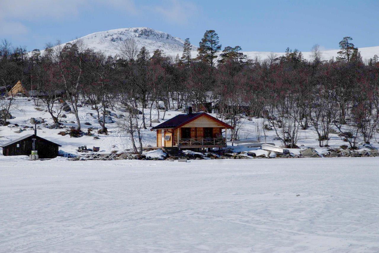 En stuga vid en frusen sjö med fjäll i bakgrunden.